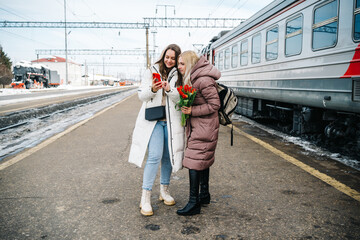 two girls on the station platform with flowers happily looking into a smartphone