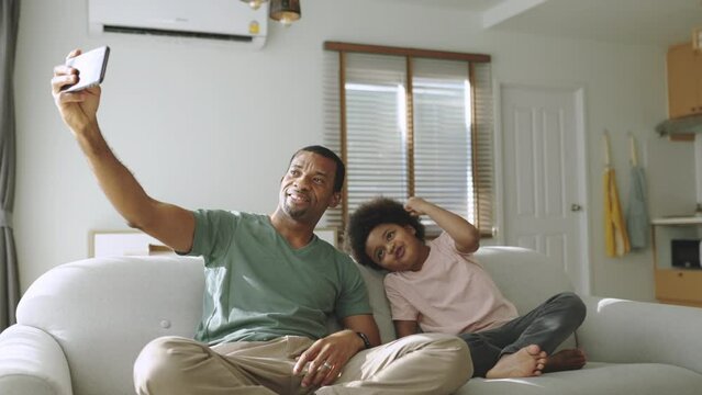 Smiling African American father and his cute boy using smartphone at home, African family doing video call on mobile phone, Black Dad and son sitting on sofa taking selfies.