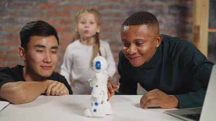 Interested man with positive Asian colleague look at moving robot on table while schoolgirl dances in spacious studio - Powered by Adobe