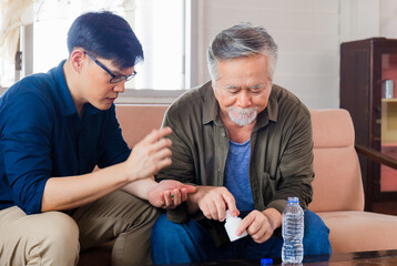 Senior father taking medication pill putting medicine on hand sitting with son in living room