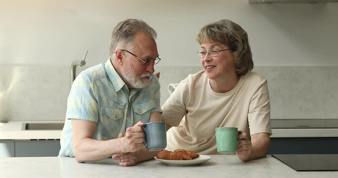 Older couple having breakfast enjoy conversation, drink fresh-brewed coffee favourite beverage, eat delicious croissants sit in kitchen at home in morning. Communication, carefree retired life concept
