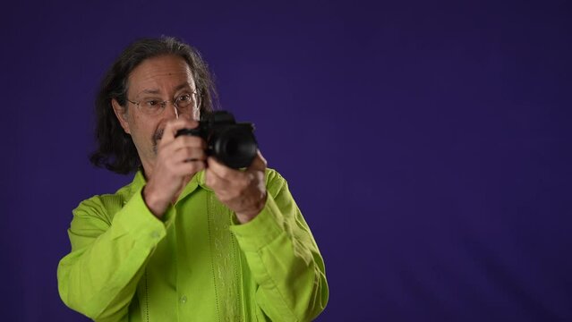 Smiling bearded man 50s 60s green shirt point finger taking photos with camera gesture isolated on solid purple background studio portrait with copy space