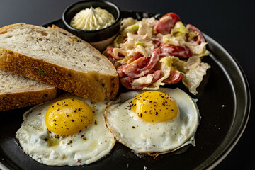 Breakfast, fried eggs on a black plate with tomatoes and chili peppers with bread on a black background