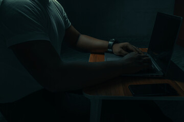 A man in a white shirt sits on a laptop in a dark room. with light shining down. internet concept...