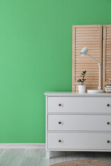 Chest of drawers with white lamp, flowerpot and folding screen near green wall