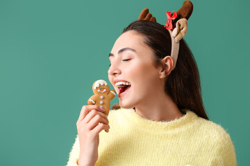 Young woman in deer horns eating gingerbread cookie on green background