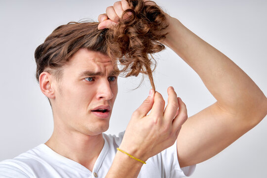 amazed confused man looking at split hair ends, want to cut isolated over light gray background. caucasian young male in casual wear looking embarrassed, need hair care. portrait copy space