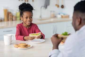 Adorable black girl looking at her dad while eating sandwich