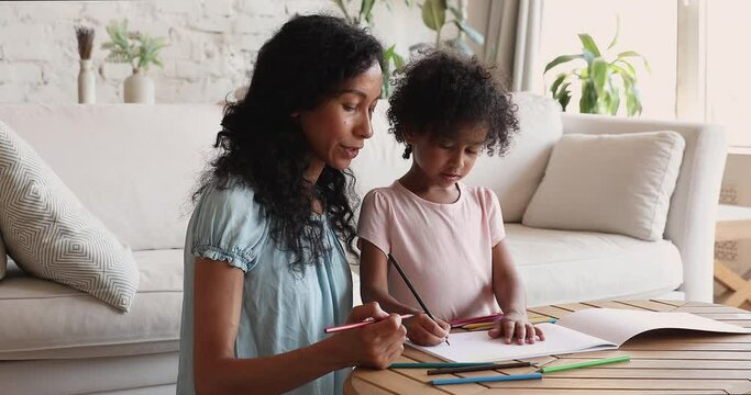 Young African mother assist 5s daughter to draw picture in sketchbook using pencil teach to make simple funny image involve in creative pastime. Happy little girl learn to make sketches imitate mom