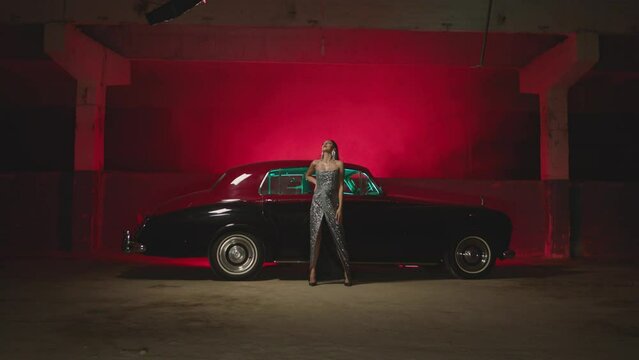 Beautiful Elegant Young Woman Standing , Dancing and Flirting near old retro classic car. Old fashioned luxury style concept . Rich fashion lifestyle . Luxury dress with fabric glitter, spangles .