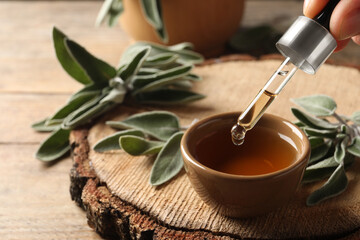 Woman dropping essential sage oil into bowl at wooden table, closeup. Space for text