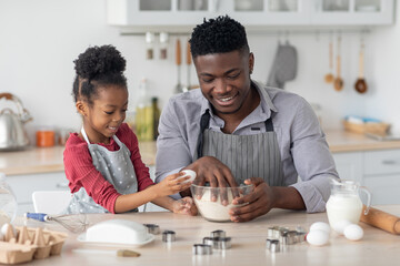 Little black girl adding eggs while her daddy making dough