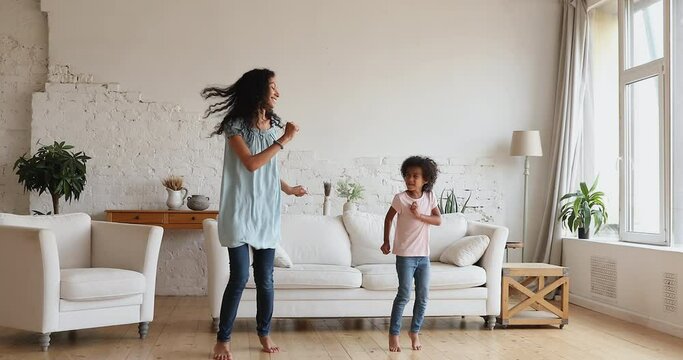 Cute active Black family two female generations young mom small daughter jump dance at living room with underfloor heating enjoy weekend. Excited nanny and little girl dancers having fun at cozy home