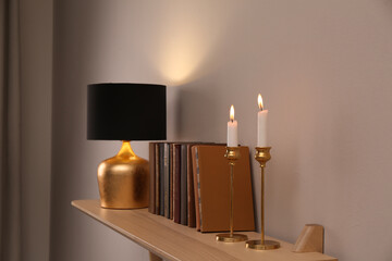 Wooden shelf with different books, burning candles and lamp on light wall