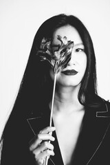 Black and white studio shot of beautiful Asian woman portrait with long black hair, black suit and makeup. Model cover her one eye with flower blossom. Flower is in camera focus