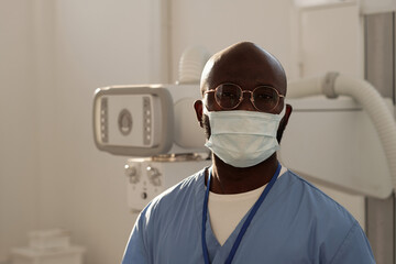 Young confident African American male assistant in uniform, eyeglasses and protective mask standing against roentgen equipment