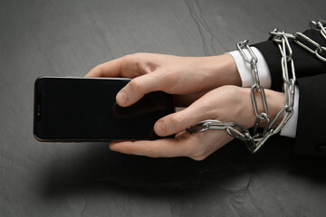 Man holding smartphone in chained hands at black table, closeup. Internet addiction