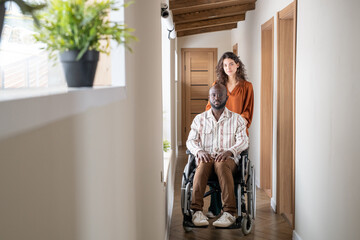 Young contemporary interracial couple standing in corridor of apartment while female caregiver standing in front of man in wheelchair