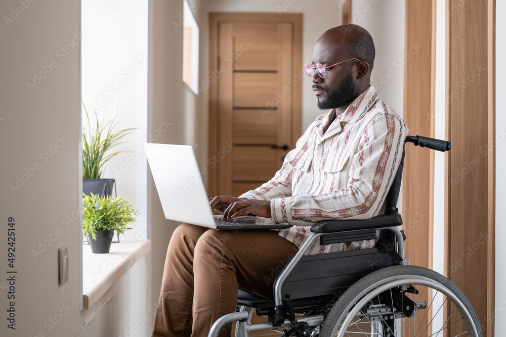 Poster young serious black man with disability concentrating on network while sitting in wheelchair in corr