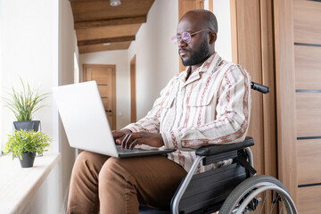 Contemporary young serious black man with laptop on his knees sitting in wheelchair and working in the net at home