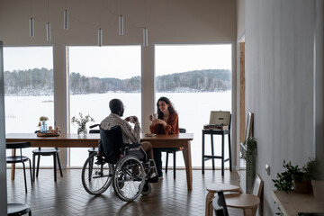 Happy young interracial couple talking and having tea by table in living room or spacious kitchen of modern apartment