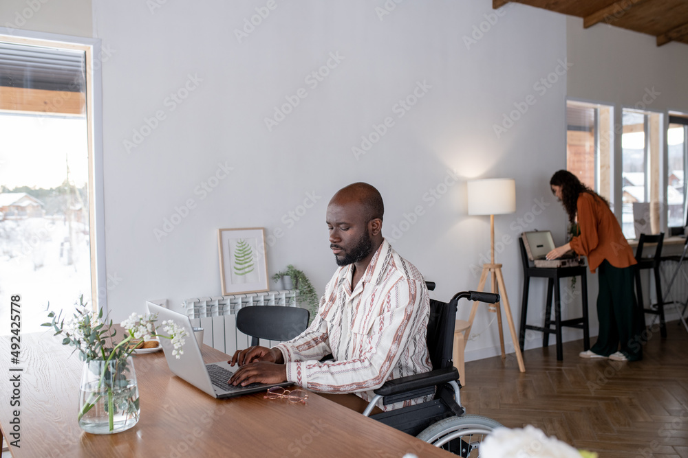 Poster serious african american man in wheelchair sitting by table in front of laptop and networking agains