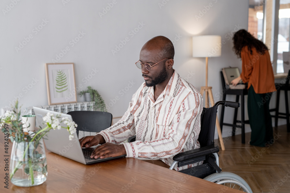 Poster young man with disability in casual shirt networking in front of laptop while sitting by table again