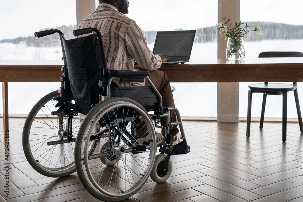 Poster back view of black man in casualwear typing on laptop keyboard while sitting by table at home and ta