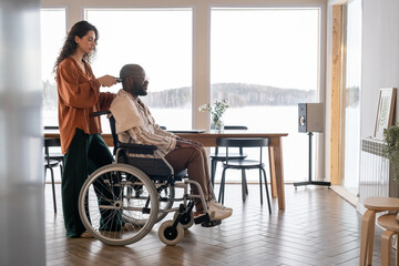 Young woman with electric trimmer cutting hair on head of man with disability sitting in wheelchair in spacious living room - obrazy, fototapety, plakaty
