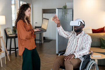 Young woman with laptop standing in front of man with disability sitting in wheelchair with vr...
