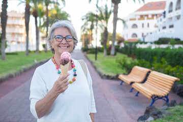 Fototapeta premium Senior woman smiling eating ice cream of chocolate and strawberry looking at camera. Happy female retiree enjoying sweet food in public park