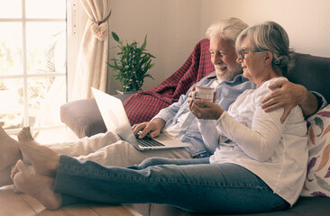 Relaxed senior couple lying on sofa at home using laptop. Two elderly retirees enjoying free time and technological devices, woman holding in hand a cup of coffee or tea
