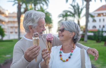 Fototapeta premium Close-up on two melting ice cream cones, held in the hand by a cheerful senior couple. Elderly people smiling looking each other in the eyes enjoying vacation or retirement