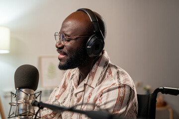 Young bald African American man in eyeglasses and headphones singing in microphone while recording...