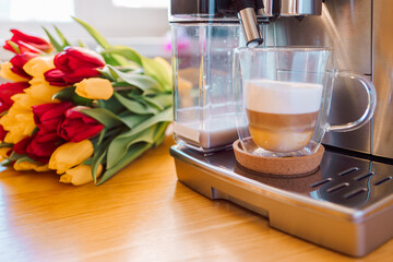 Cup of coffee with milk and tulips flowers on wooden kitchen table. Freshly brewed cappuccino espresso in coffee machine for breakfast. Stylish home interior decor. High quality photo
