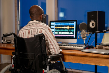 Rear view of man with disability using sound recording program while sitting by table in front of computer monitor in studio