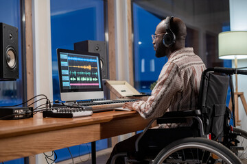 Young serious African American musician with disability looking at computer screen with sound visualization in studio - obrazy, fototapety, plakaty