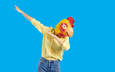 Funny girl disguised as hen or chicken dancing in the studio. Portrait of young woman wearing strange absurd surreal eccentric wacky carnival rooster mask dabbing isolated on solid blue background