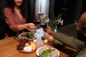 Young amorous interracial couple toasting with red wine over festive table served with healthy food and candles for romantic dinner