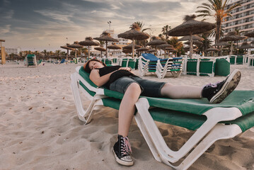 Young woman with long red and black colored hair lying down on a deckchair at the beach, looking at...