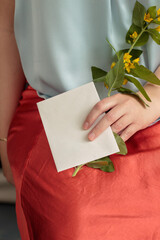 Woman's hand holding blank envelope and flowers closeup with orange skirt and blue shirt.