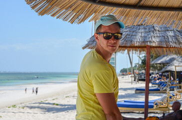 Man in a cap and sunglasses standing on the beach under an umbrella, Diani Beach, Kenya, Africa
