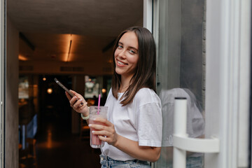 Happy smiling girl with wonderful smile with dark hair wearing white t-shirt is using smartphone and drinking smoothie while is leaving cafe 