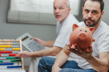 Man showing a piggy bank while his senior father is calculating