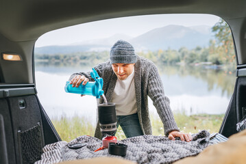 A smiling man dressed in warm knitted clothes pouring a freshwater in gas burner stove in the cozy car trunk with a beautiful mountain lake view. Warm early autumn auto traveling concept image