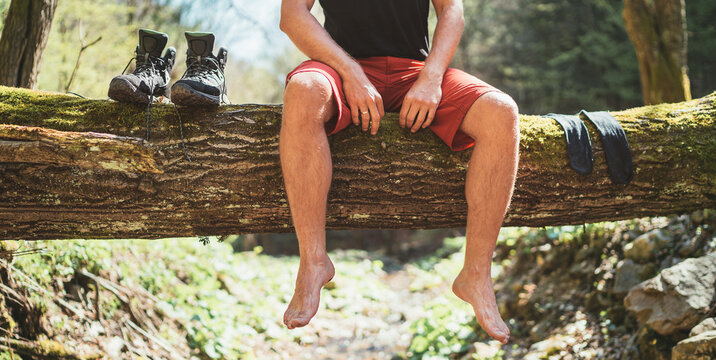  Man sitting on a fallen tree log over the mountain forest river while he waiting for socks nd trekking boots laundry drying. Active people traveling, hiking or trekking concept image.