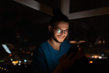 Night portrait of smiling man looking in smartphone on background of window.