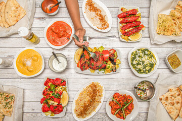 Indian food dishes, with fried chicken tikka masala, garlic naan, yogurt cream, korma, tandoori chicken, tomato salad, palak paneer, pepper, basmati rice and hand with fork stabbing the chicken