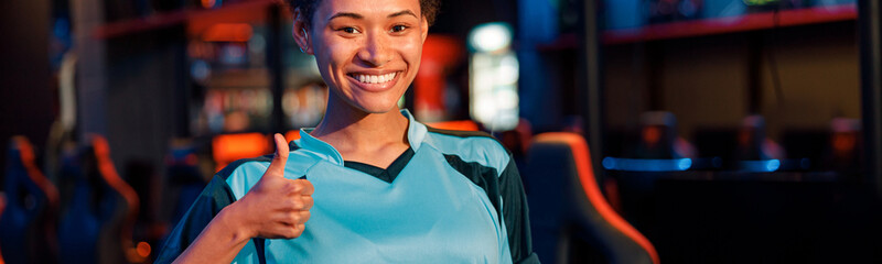 Young female esports gamer giving thumbs-up sign with cheerful facial expression showing success at...