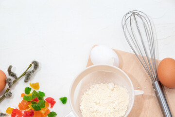 Sieve with flour on wooden board, eggs, candied fruits, whisk and sprig of willow. Background for food advertising, Easter with place for text. Selective focus.